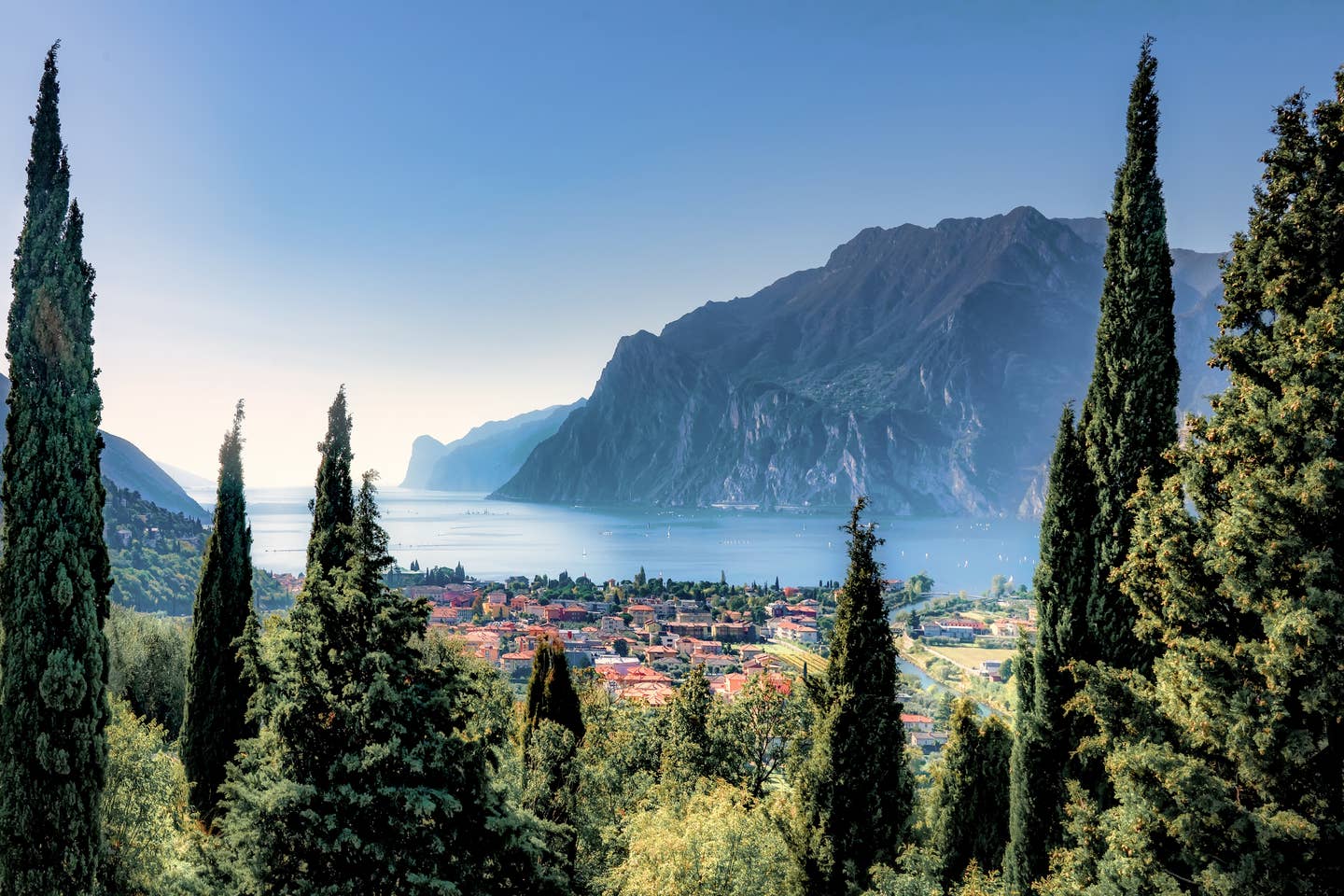 Gardasee Urlaub mit DERTOUR. Von Zypressen eingerahmter Blick auf den Ort Torbole mit dem Gardasee und Bergen im Hintergrund