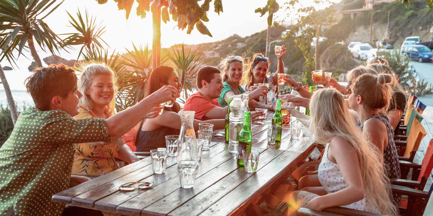 Gruppe von Freunden feiert in einer Taverne am Strand mit Sonnenuntergang über dem Meer im Hintergrunfd