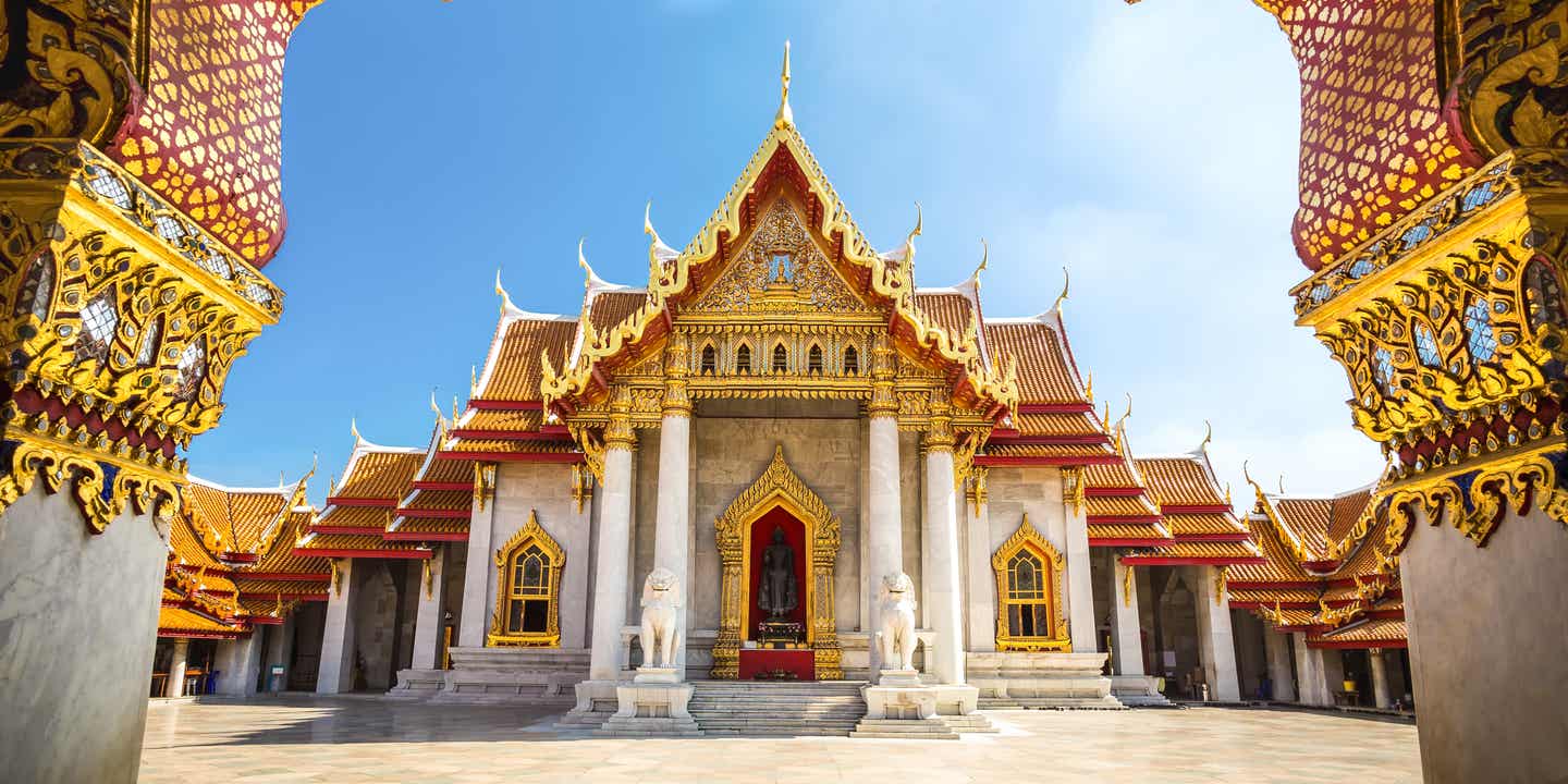 Blick durch einen verzierten Bogengang auf einen typischen thailändischen Tempel mit weißem Marmor, roten Dächern und Gold-Verzierungen.