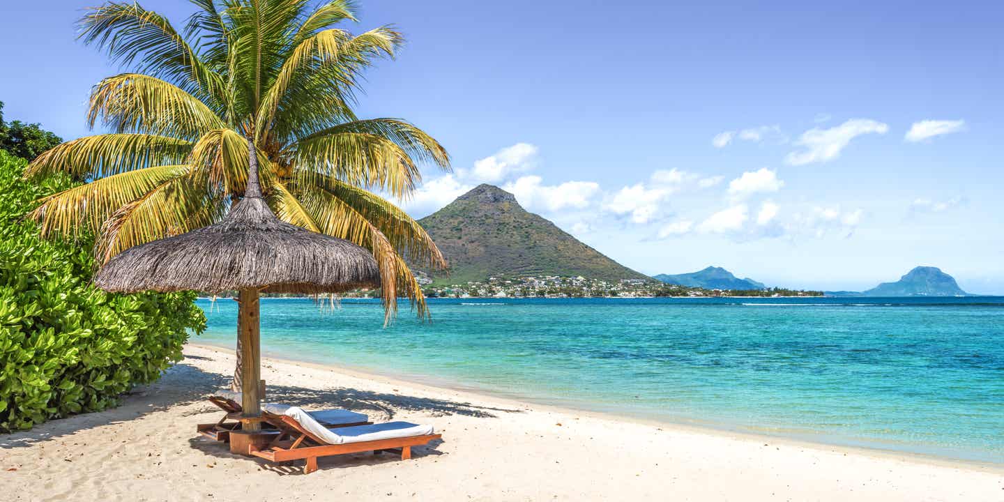 Liege und Sonnenschirm am Strand von Mauritius. Im Hintergrund Berge der Insel.