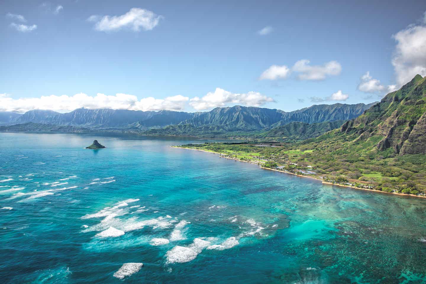 Luftaufnahme der Küste von Oahu; im Vordergrund das türkisfarbene Meer, im Hintergrund eine langgezogene Bergkette.