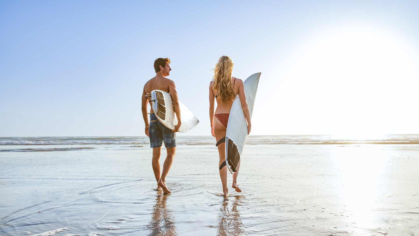 Paar mit Surfbrettern geht bei Sonnenuntergang am Sandstrand von Tarifa in Richtung Meer spazieren