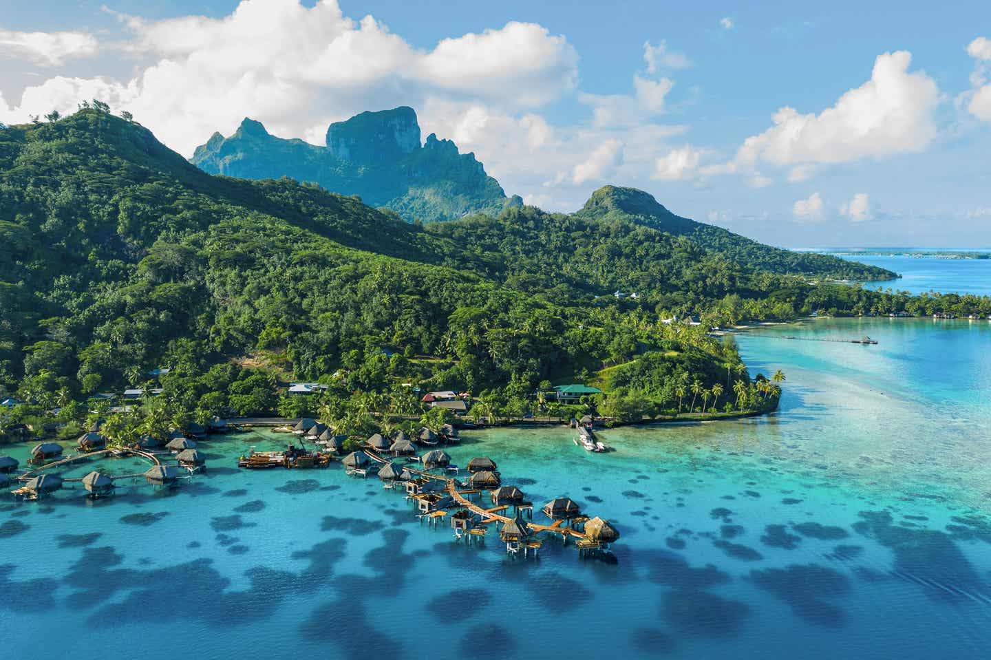 Blick auf die von grünen Wäldern gesäumte Küste von Bora Bora; im Vordergrund türkisblaues Meer, im Hintergrund die Inselberge