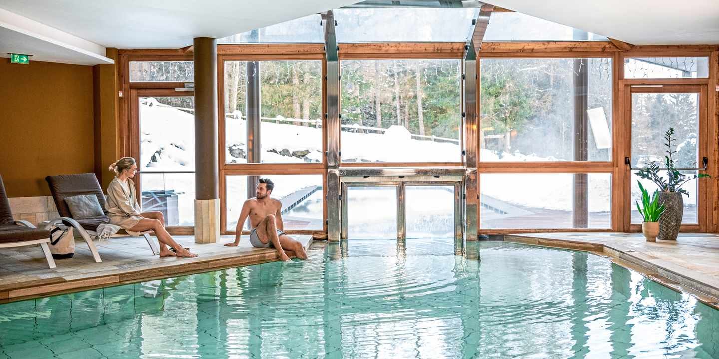 Ein Paar entspannt am Innenpool eines Wellnesshotels im Hotel Falkensteiner in Trentino-Südtirol mit Blick auf die verschneite Berglandschaft