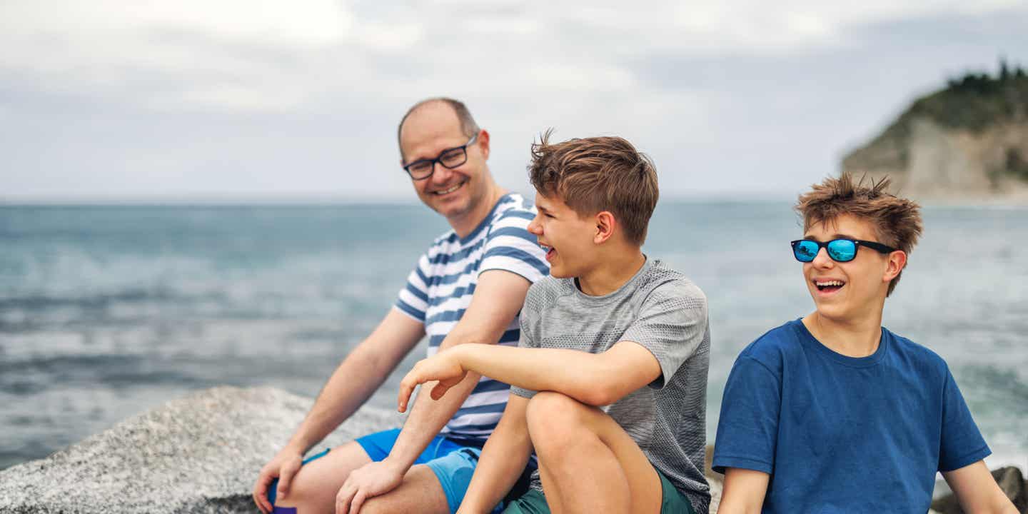 Vater und zwei Söhne sitzen auf Felsen am Strand, im Hintergrund das Meer und eine bewachsene Klippe.