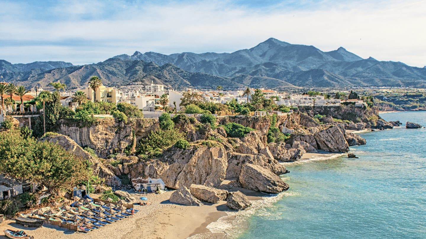 Andalusien Sehenswürdigkeit: Kleine Badebucht in Nerja mit Liegestühlen, Felsen, Altstadtblick und den Bergen der Axarquía im Hintergrund 