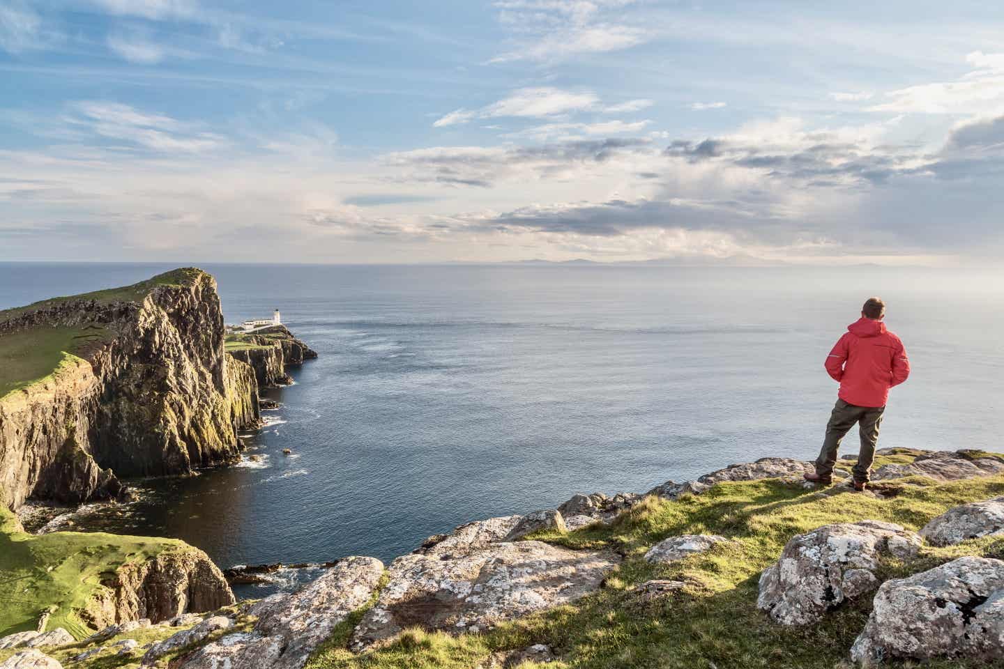 Ein Mann mit roter Jacke steht auf einer Klippe am neist Point der Isle of Skye und schaut auf einen Leuchtturm in der Ferne