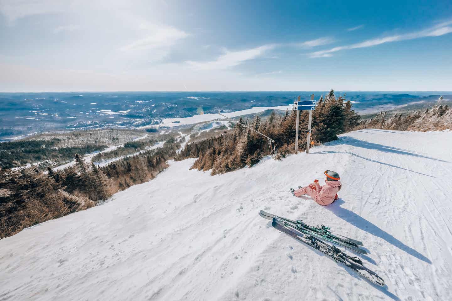 Eine Skifahrerin sitzt im Schnee und genießt vom Berg aus den Blick in das verschneite Tal und die Weiten Kanadas.