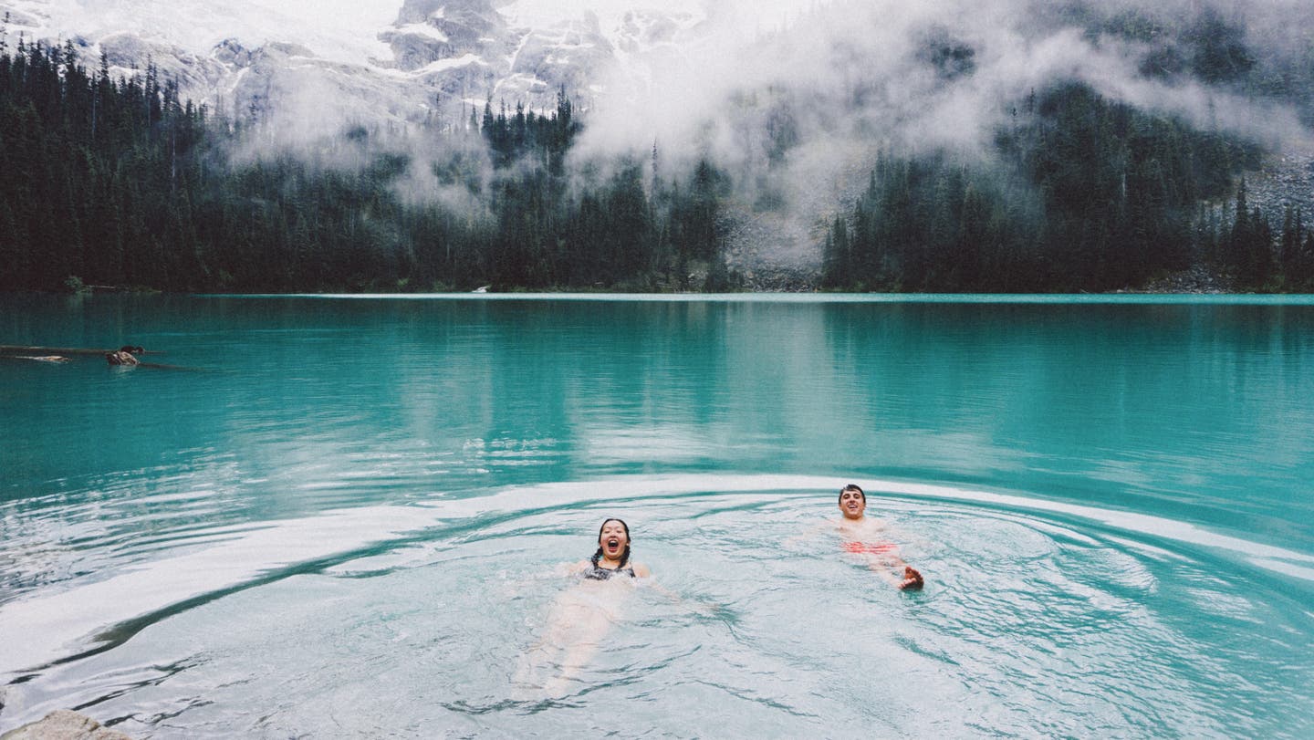 Paar macht Eisbaden in blauem See vor Bergen und Tannen