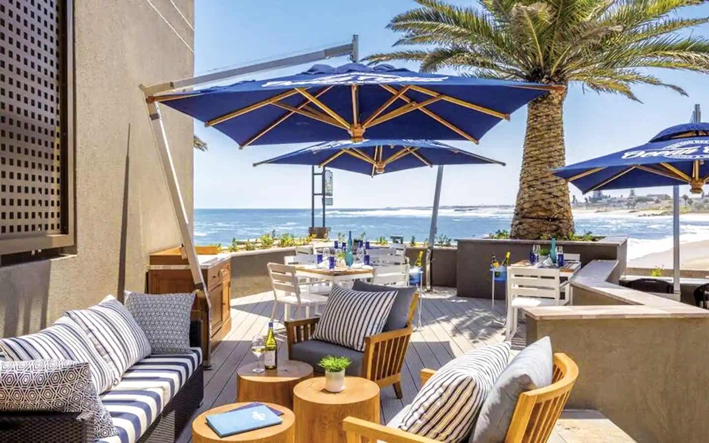 Terrasse des Strand Hotel in Swakopmund in Namibia mit gemütlicher Lounge, Meerblick, Palmen und Sonnenschirmen direkt am Atlantik Sundowner mit Meerblick im Strand Hotel in Swakopmund