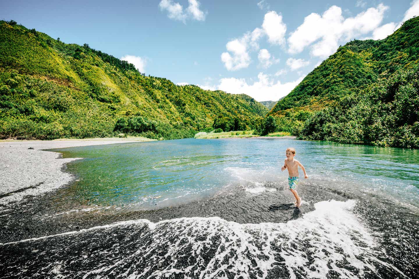 Ein kleiner Junge spielt in einem natürlichen Wasserbecken auf Maui, im Hintergrund üppige grüne Wälder