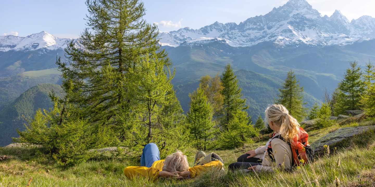 Zwei Freundinnen entspannen auf einer Bergwiese mit Blick auf schneebedeckte Gipfel während eines Freundinnen Wellnesswochenendes