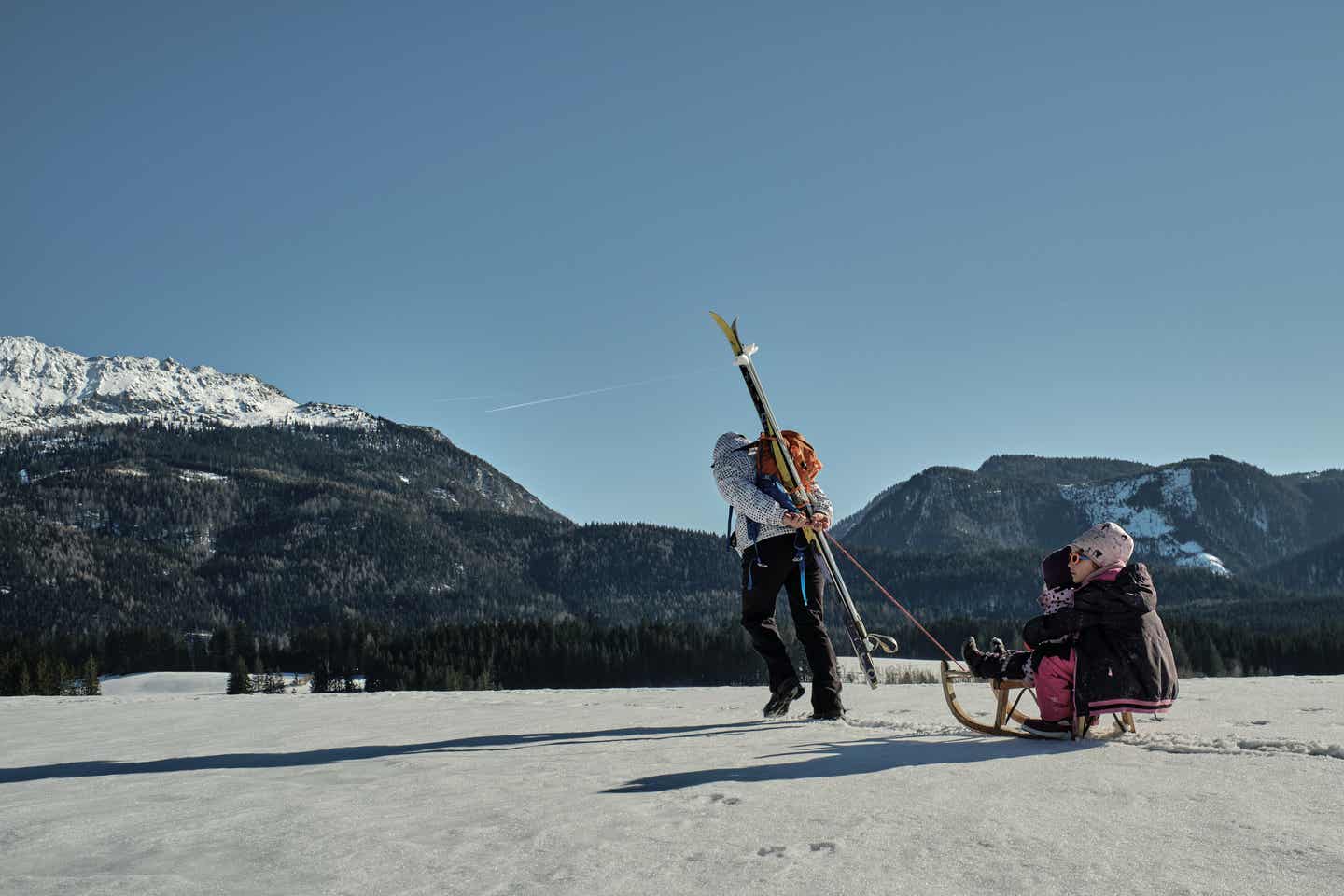 Österreich Urlaub mit DERTOUR. Mutter zieht ihre Kinder auf einem Schlitten durch eine sonnige Winterlandschaft in der Steiermark