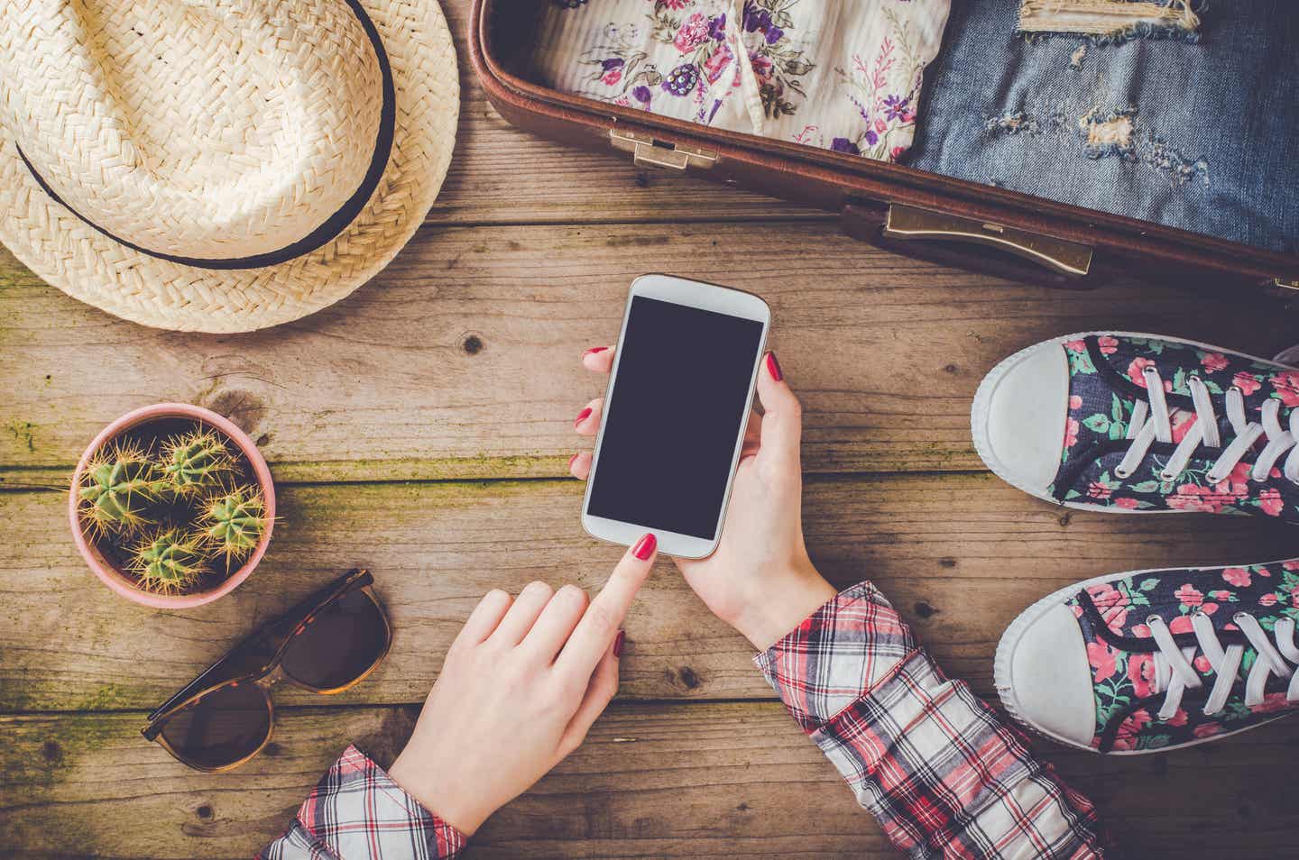 Nachhaltigere Reisevorbereitungen mit Apps: Frau plant Urlaub per Smartphone neben Koffer, Hut und nachhaltigem Gepäck
