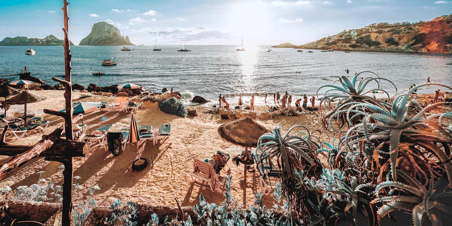 Blick auf Cala d’Hort auf Ibiza mit Es Vedrà im Sonnenlicht und Urlaubsgästen am Strand
