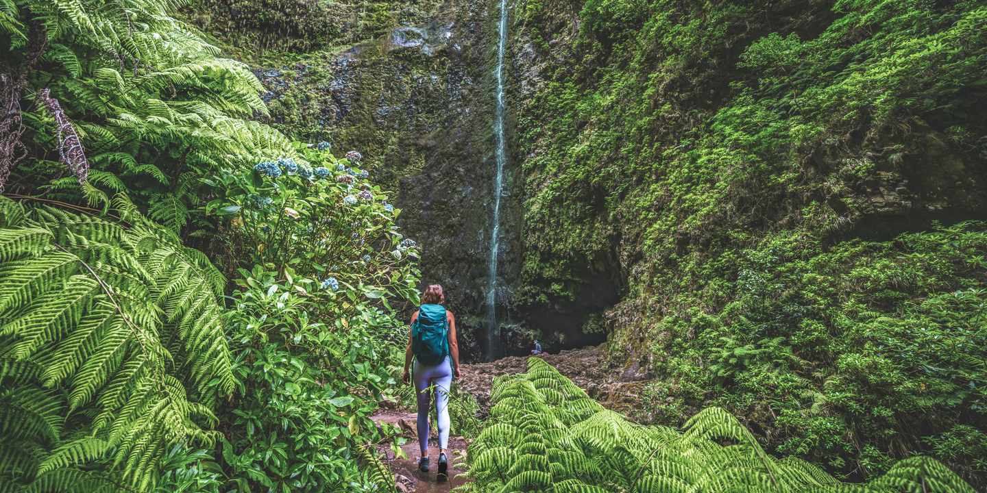 Wanderin mit Rucksack auf schmalem Pfad zur Levada Caldeirão Verde vor hohem Wasserfall