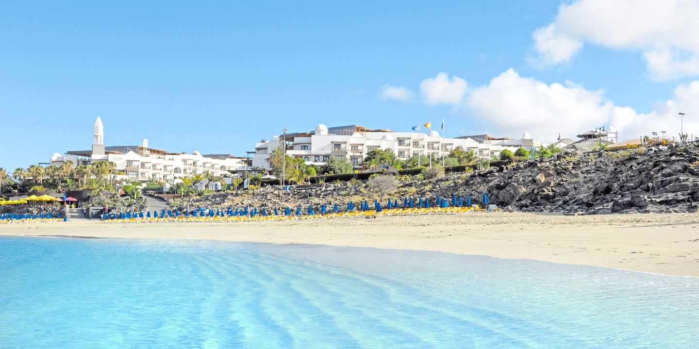 Blick vom Meer aus auf den Strand mit Klippen und weißem Hotel oben auf
