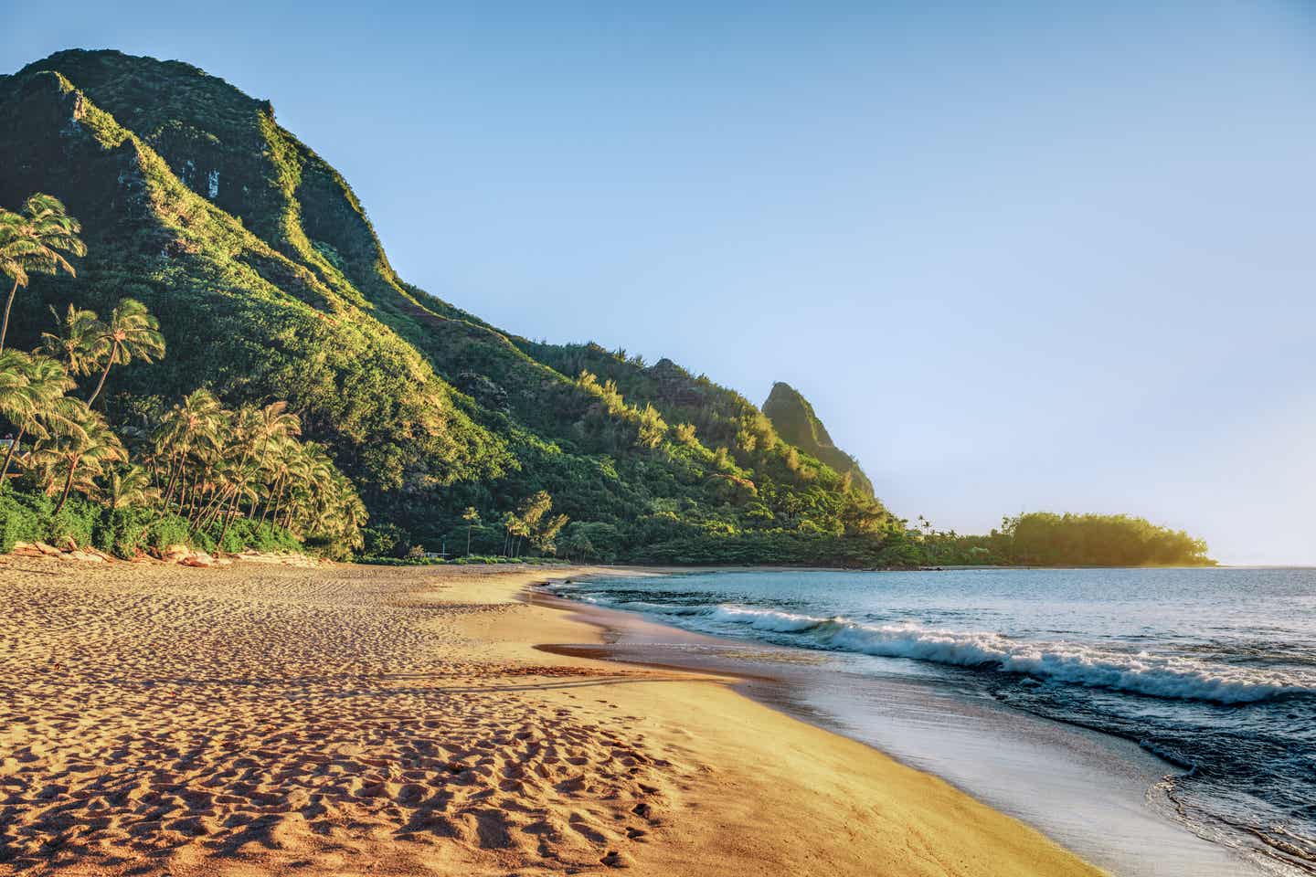 Ein Strand auf Kauai im Licht der untergehenden Sonne; links der Sandstrand, rechts das schimmernde Meer und im Hintergrund Palmen und ein grün bewachsener Hügel