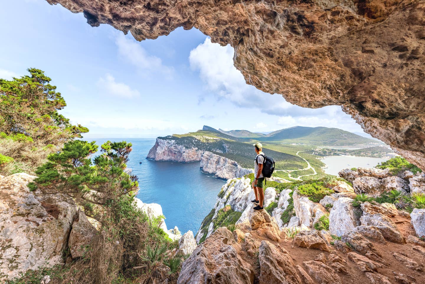 Wanderer mit Rucksack blickt von einer Höhle bei Capo Caccia auf das Meer und die Steilküste Sardiniens Wanderer mit Rucksack blickt von einer Höhle bei Capo Caccia auf das Meer und die Steilküste Sardiniens