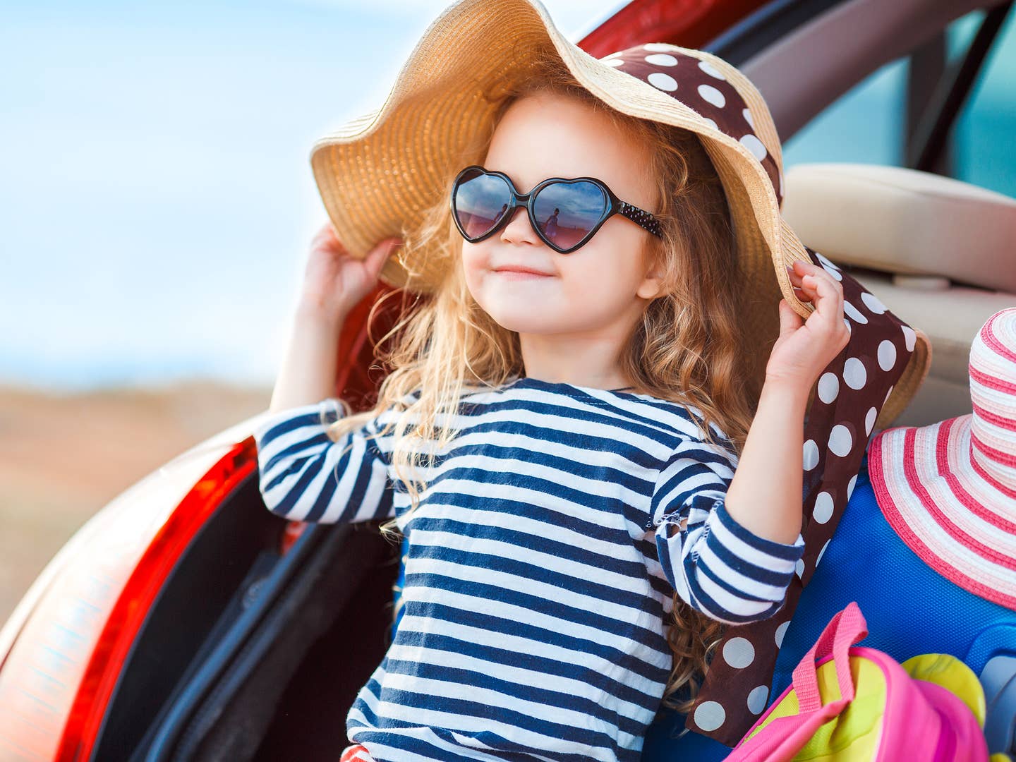 Auf Rundreisen für Familien: Kleines Mädchen mit Sonnenhut und Sonnenbrille sitzt im Kofferraum eines Autos Auf Rundreisen für Familien: Kleines Mädchen mit Sonnenhut und Sonnenbrille sitzt im Kofferraum eines Autos