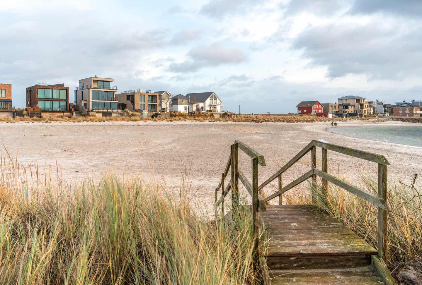 Holzsteg führt durch Dünengras zum Sandstrand in Olpenitz an der Ostsee, mit modernen Ferienhäusern und Blick auf das Meer