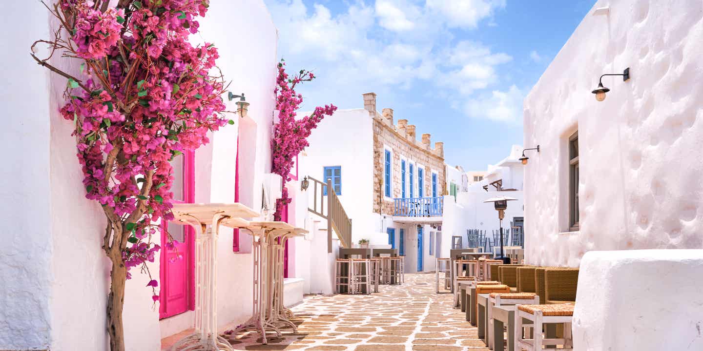 Gasse in Naoussa auf Paros mit weißen Häusern, pinker Bougainville und blauen Fenstern unter Sommerhimmel