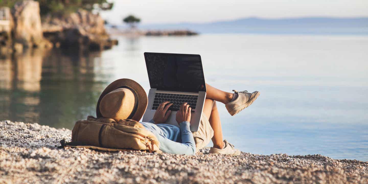 Frau liegt entspannt am Kiesstrand und plant ihre nachhaltige Reise und Ausflüge mit einem Laptop am Meer
