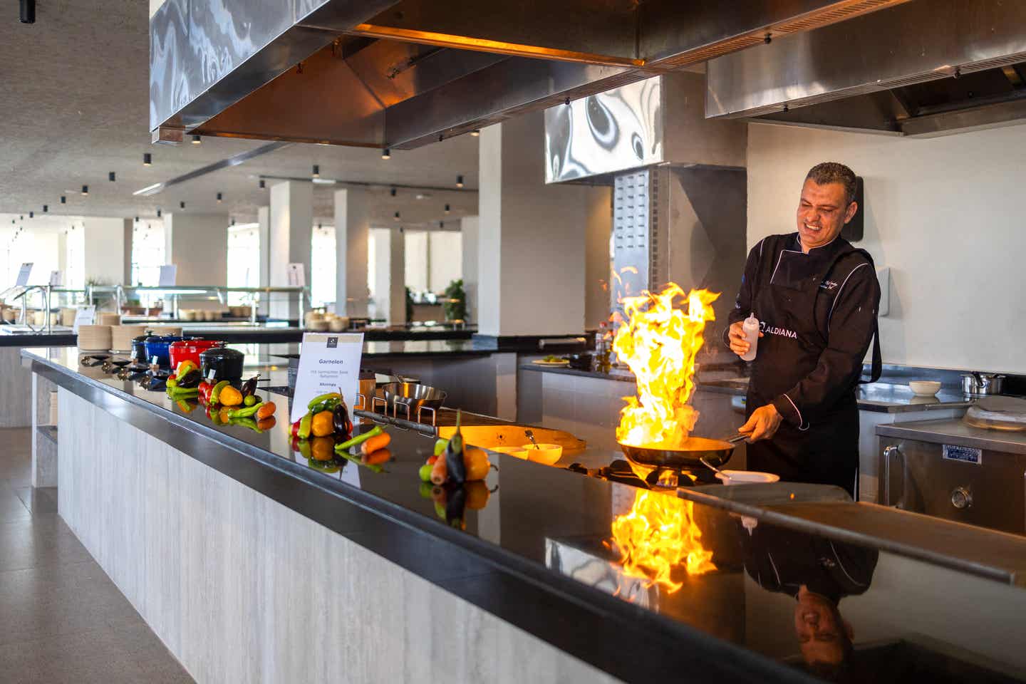 Koch im Aldiana Club Hotel Naga Bay flambiert Speisen an einer offenen Show-Cooking-Station mit frischem Gemüse im Vordergrund