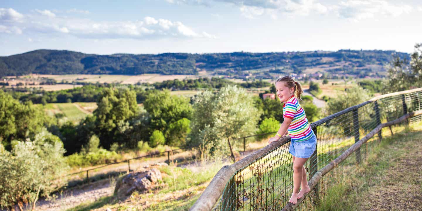Kind lehnt auf einem Geländer und blickt auf eine hügelige Landschaft mit Bäumen und Feldern.