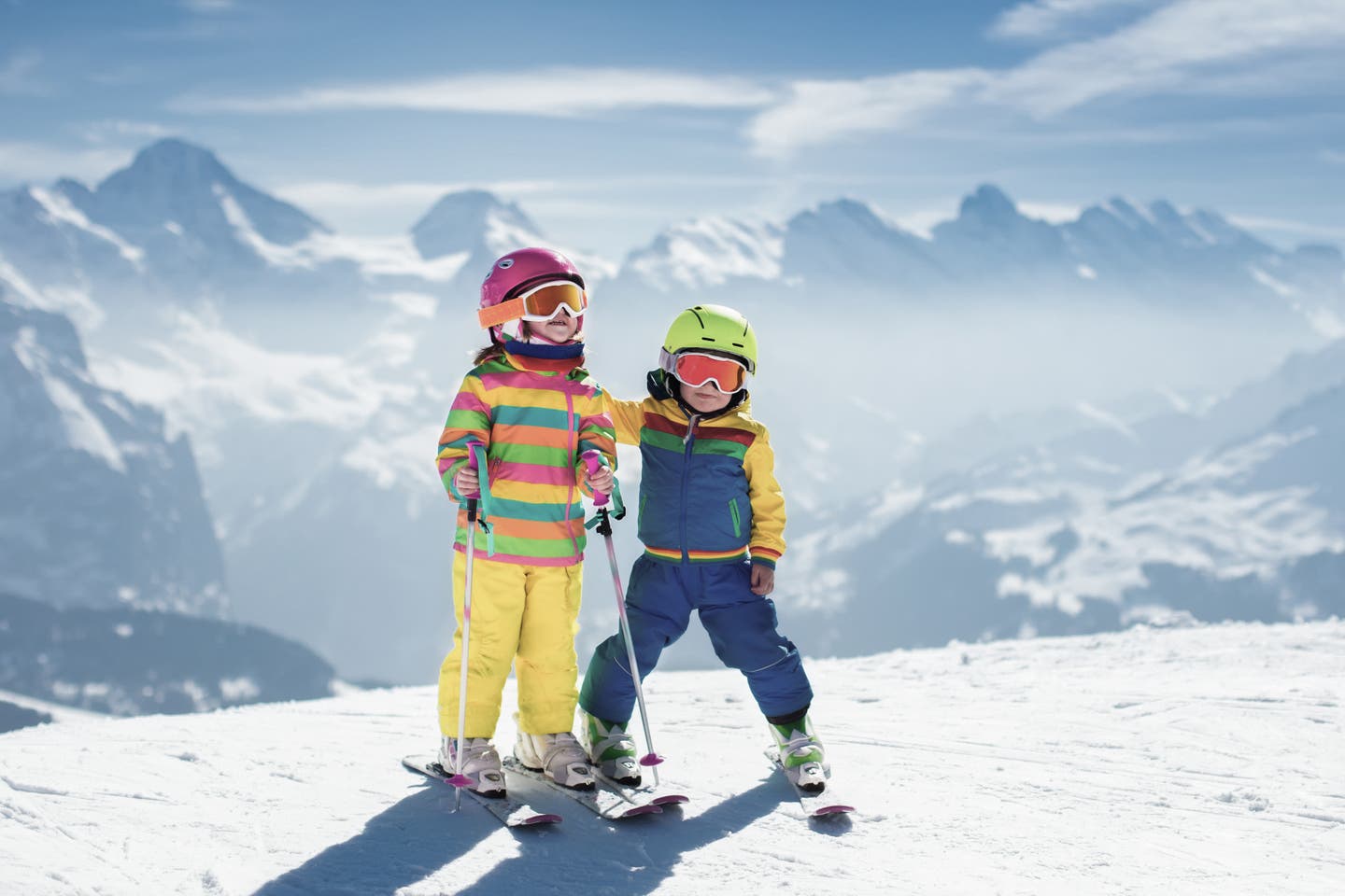 Zwei Kindern mit Skiern auf einer Piste vor Bergen