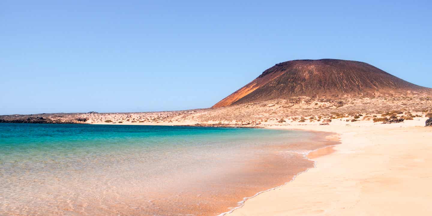 Rosafarbener Sand vor blauem Wasser an einem Strand von La Graciosa, ein Vulkan im Hintergrund
