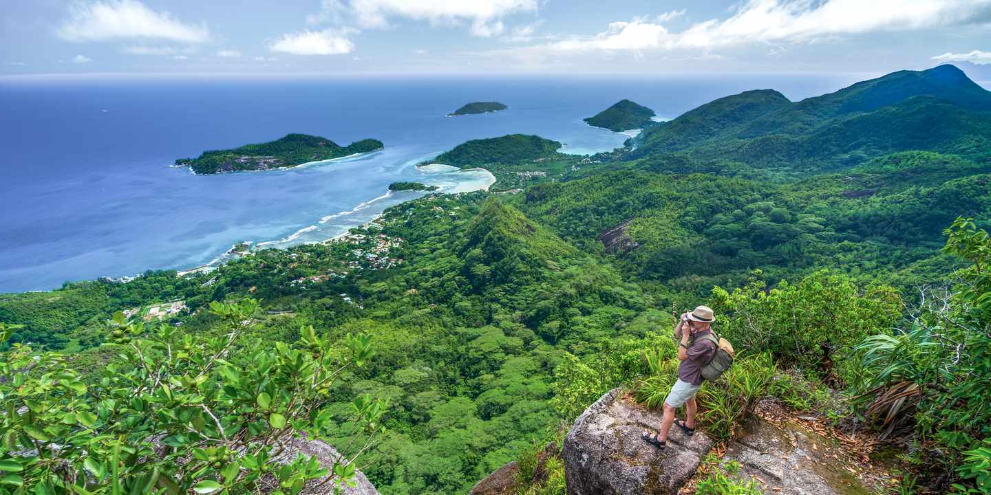 Die Füße in warmen, weichen Sand stecken und im Schatten der Palmen eine Kokosnuss schlürfen – wenn das Ein Wanderer steht auf einem Felsen auf der Insel Mahé und blickt über grüne Hügel, Buchten und den Indischen Ozean  du, was dich dort alles erwartet und wann die beste Reisezeit ist. 