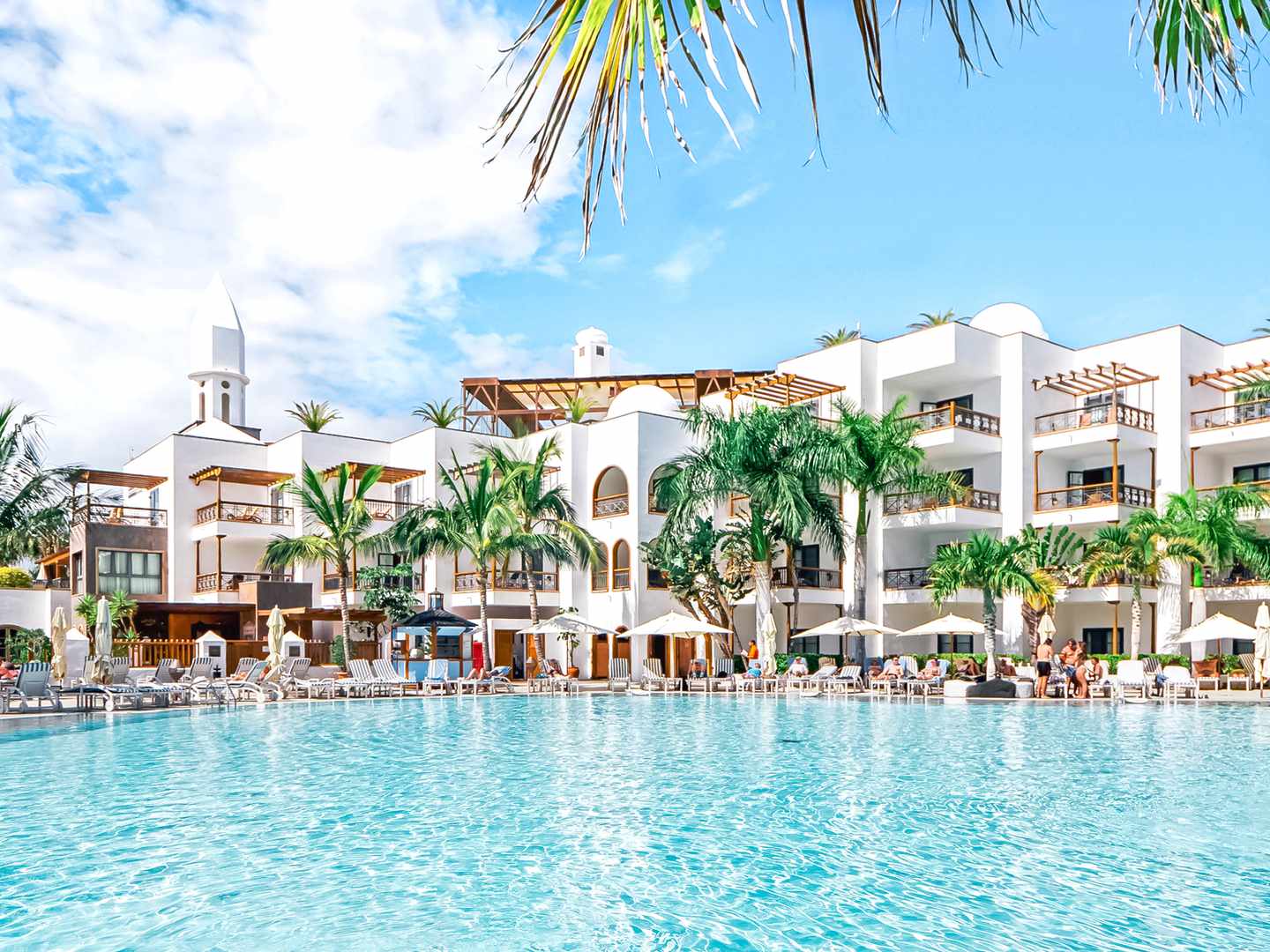 Eine traumhafte Anlage für ein Wellnesshotels auf Lanzarote Poolbereich des Hotels mit dahinterliegendem weißen Gebäude und grünen Palmen