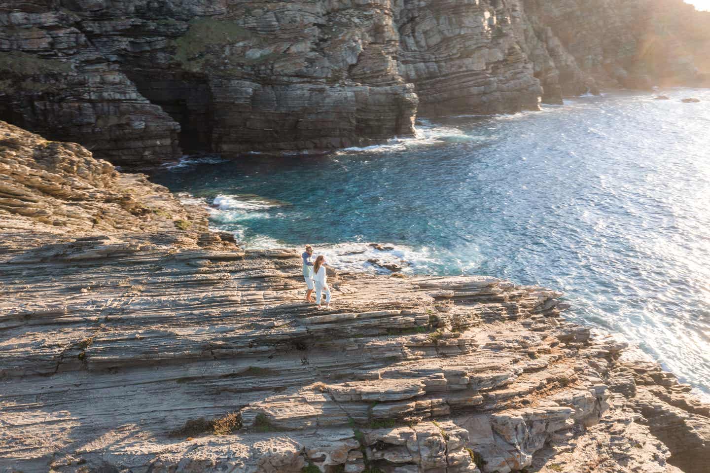 Sardinien Urlaub mit DERTOUR. Luftaufnahme eines Paares in weißen Kleidern auf einer Klippe am Meer vor Sardinien