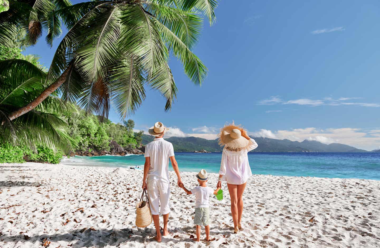 Familie mit Mutter, Vater und Kind im Mahé-Urlaub am hellen Sandstrand, gesäumt von Palmen und Bergen