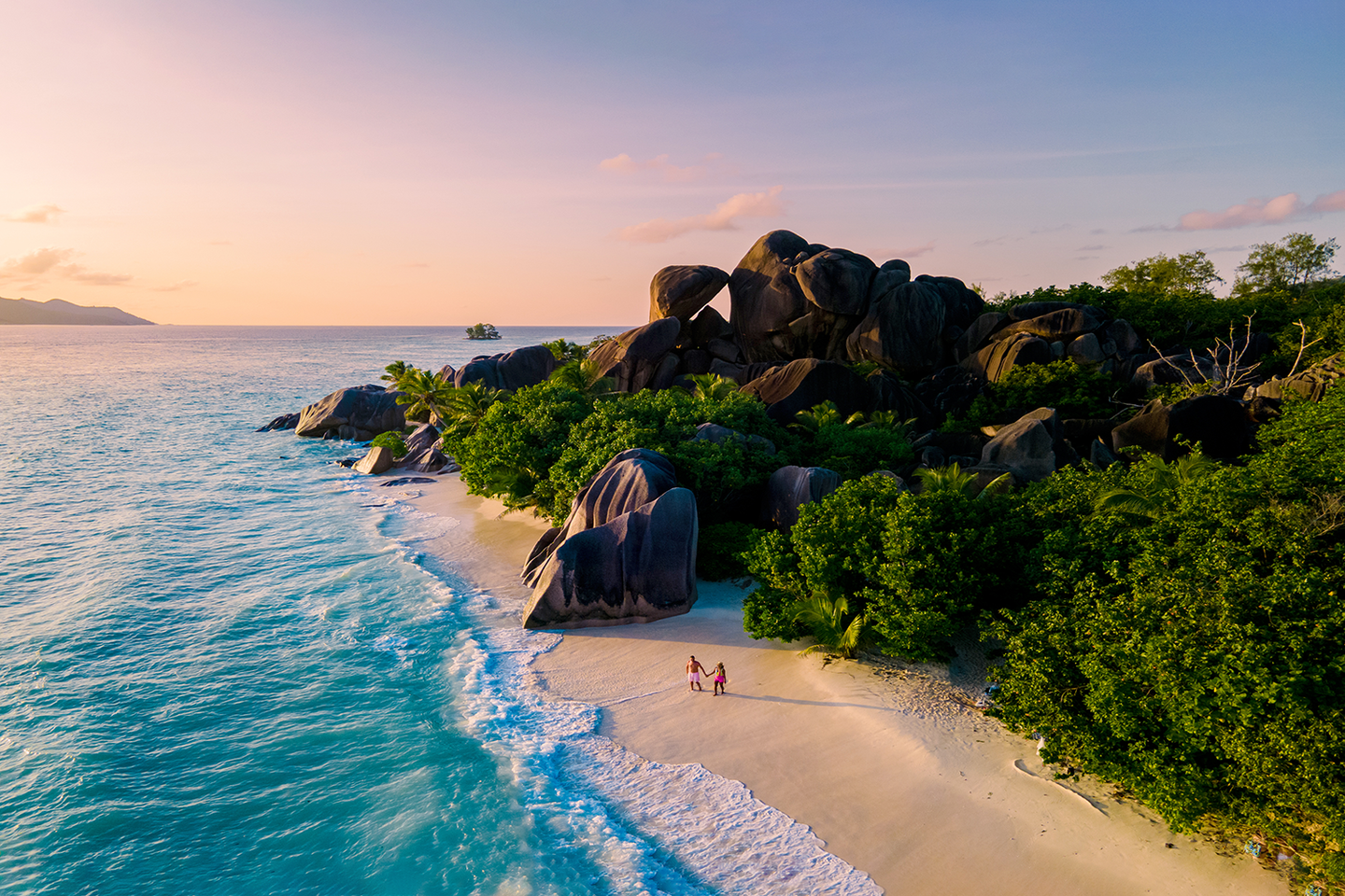 Ein Paar macht Urlaub im Indischen Ozean und spaziert Hand in Hand am Strand Anse Source d’Argent auf den Seychellen bei Sonnenuntergang, im Hintergrund Felsen und tropisches Grün