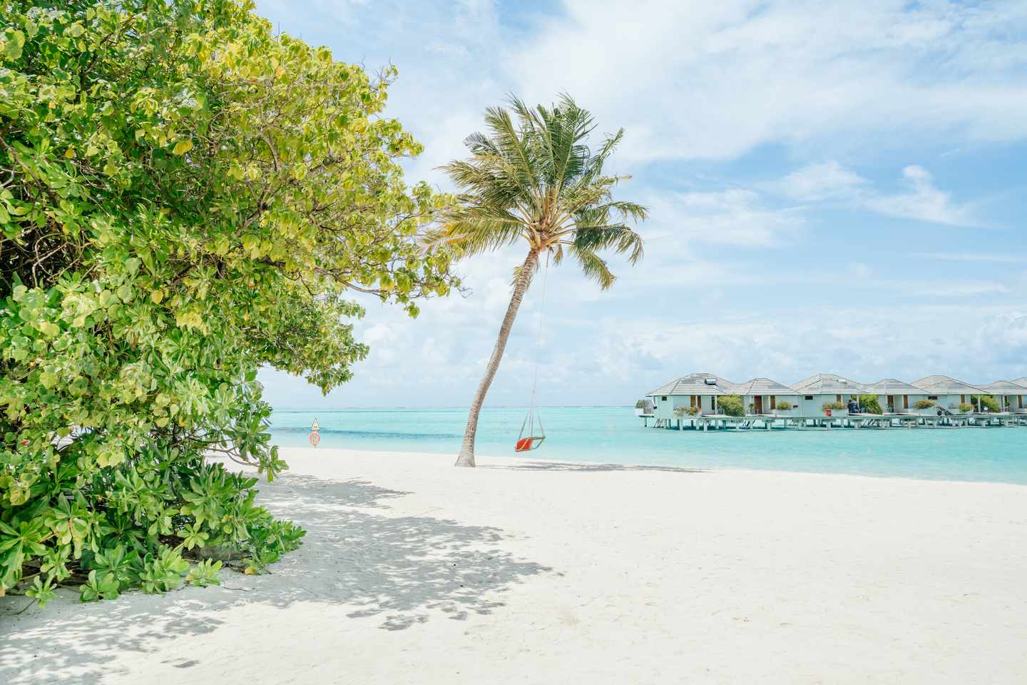 Strand mit weißem Sand, einer Palme mit einer Hängematte und über dem Wasser gebauten Villen im Hintergrund