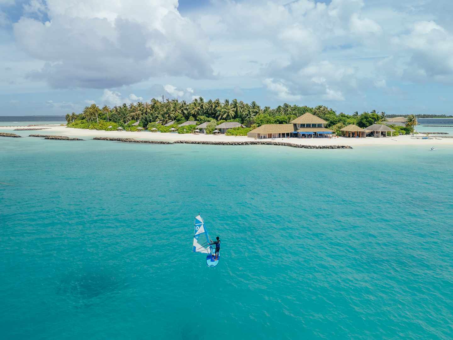 Person auf einem Windsurfbrett im türkisfarbenen Wasser, die auf tropische Insel mit Hotelanlage zusteuert