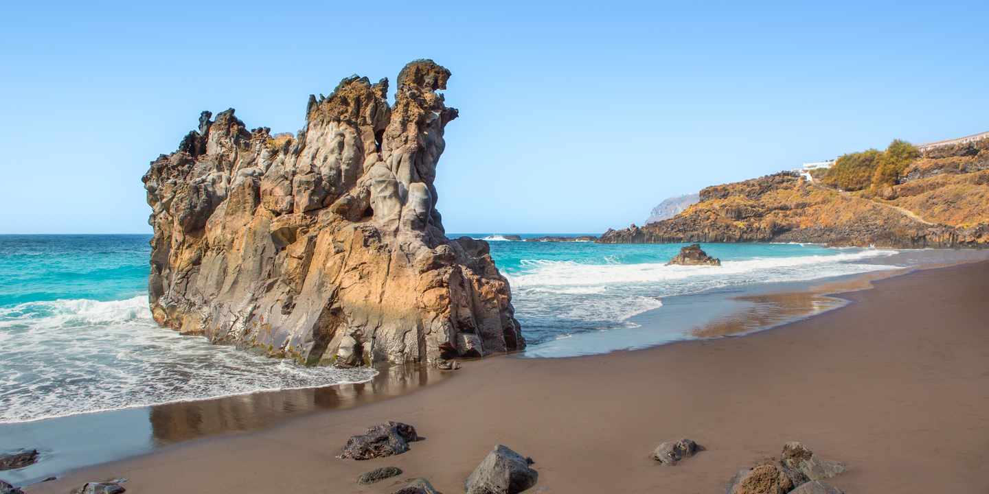 Schwarzer Sandstrand El Bollullo auf Teneriffa mit markantem Felsen im Meer und Brandung nahe Puerto de la Cruz