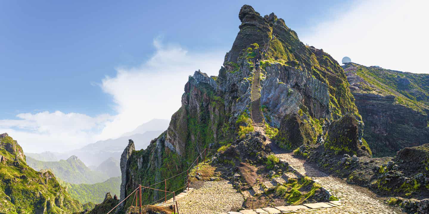 Gepflasteter Wanderweg führt durch steile Felslandschaft zum Gipfel des Pico do Arieiro auf Madeira. Helle Wolken am blauen Himmel, grüne Vegetation auf den Felsen.