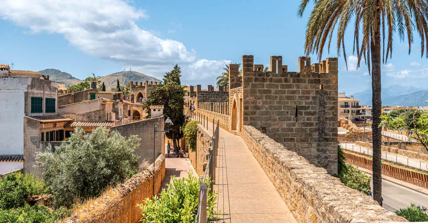 Blick auf die historischen Stadtmauern der Porta del Moll Festung, eine Mallorca-Sehenswürdigkeit, in der Altstadt von Alcúdia mit blauem Himmel und Palmen