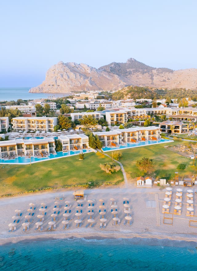 Sentido Hotels Blick auf das Sentido Port Royal auf Rhodos; im Vordergrund der Strand mit Schirmen und Liegen, im Hintergrund eine Bergkette