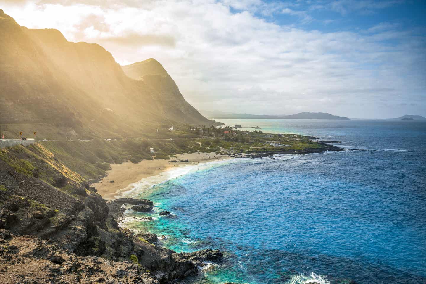 Blick auf den Makapuu Beach, einer der schönsten Hawaii-Strände, mit türkisblauem Wasser vor grün bewachsenen Bergen im Gegenlicht.