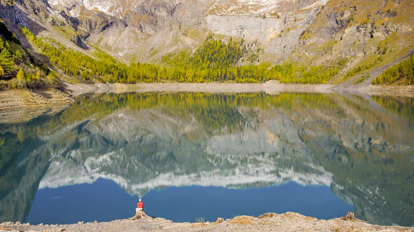Mann am Lac de Tseuzier im Wallis mit Blick auf Bergspiegelung und verschneite Gipfel der Region Crans Montana
