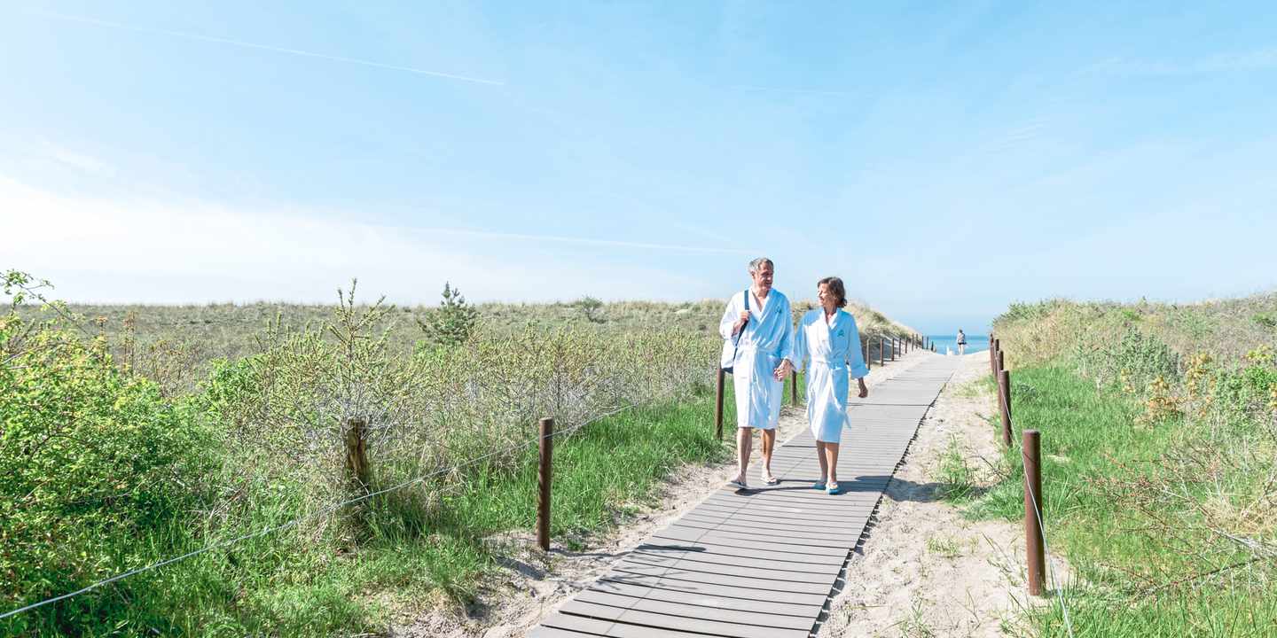 Paar im Bademantel spaziert während eines Wellnessurlaubs für Paare über den Dünenweg vom Warnemünder Ostseestrand zurück zum Neptun Hotel