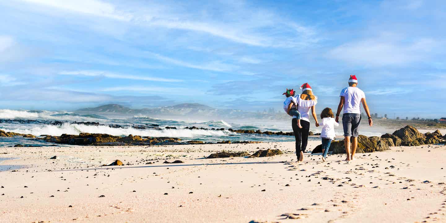 Familie mit Weihnachtsmützen aber in T-Shirts spaziert am Strand vor kräftigen Wellen