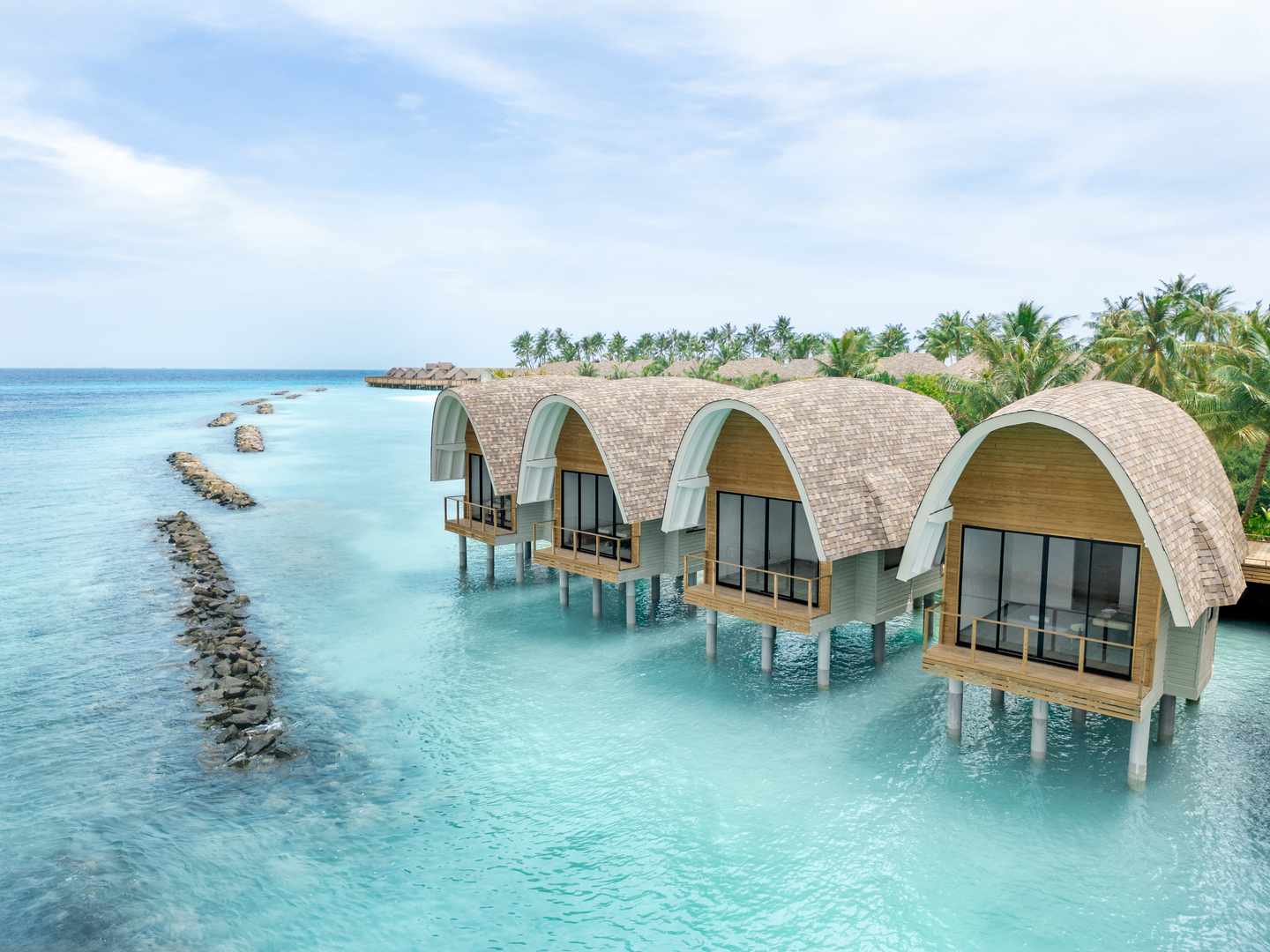 Ananea Madivaru: Vier Wasserbungalows mit ovalem Strohdach vor tropischer Insel mit Palmen