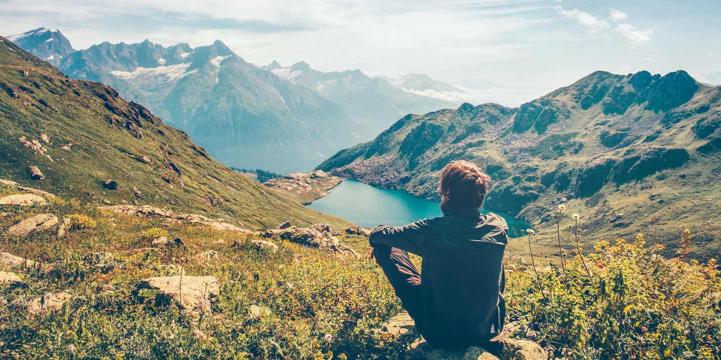 Ein Wander sitzt auf einem Stein und blickt auf einen Bergsee und die Berge im Hintergrund