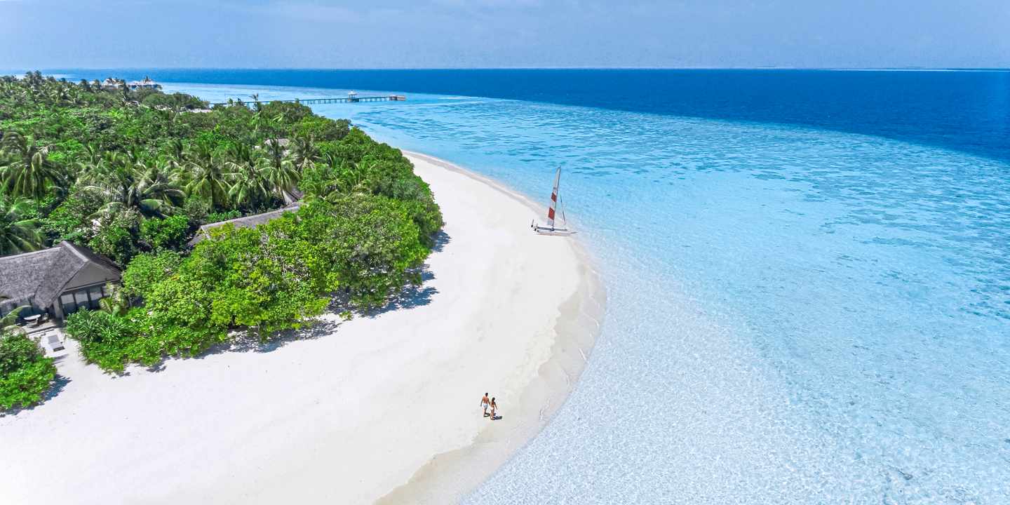Luftaufnahme eines weißen Sandstrands mit zwei Personen, umgeben von dichtem Grün und türkisfarbenem Meer im Vakkaru Maldives