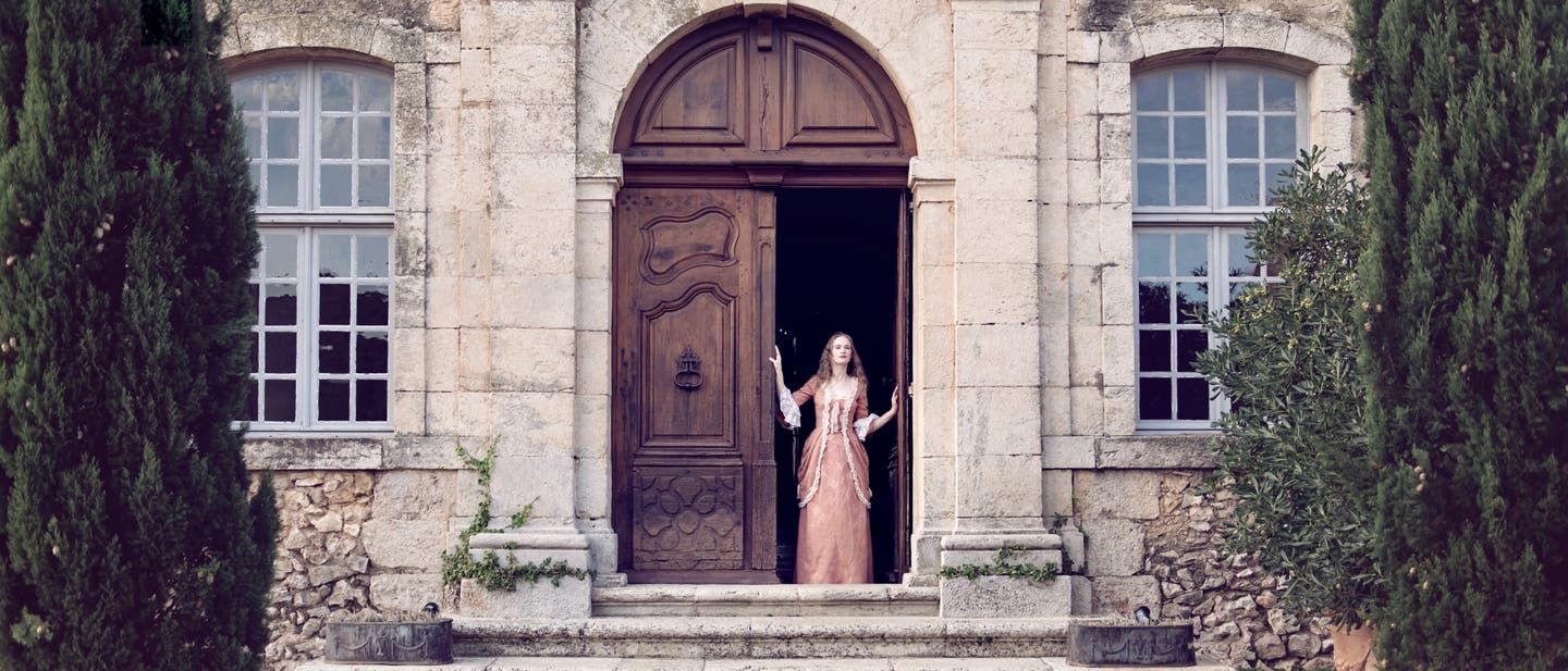 Typisch Castlecore: Eine Frau steht in einem historisch anmutenden Kleid in der alten Holztür eines steinernden Anwesens